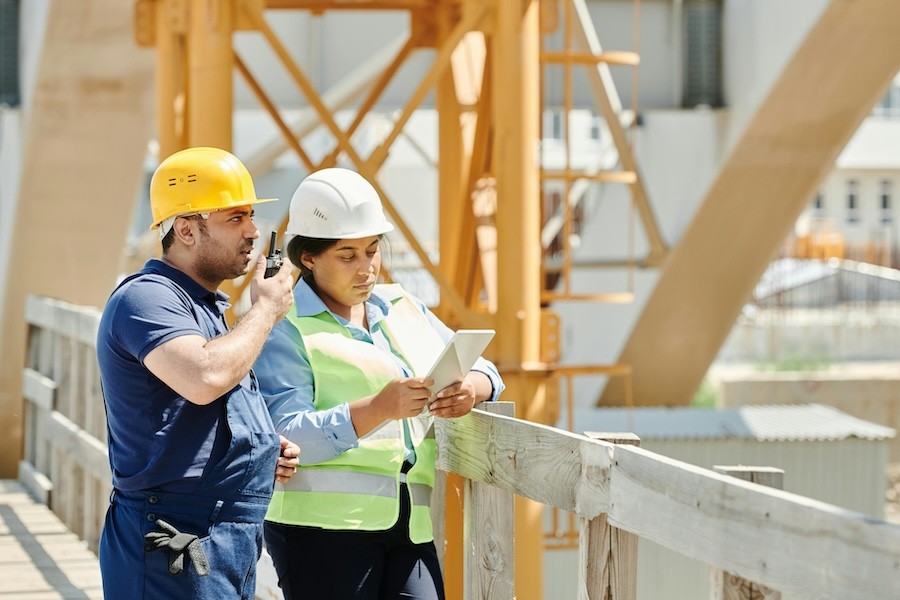 Two construction workers on a work site, one looking at a tablet and the other speaking into a walkie-talkie.