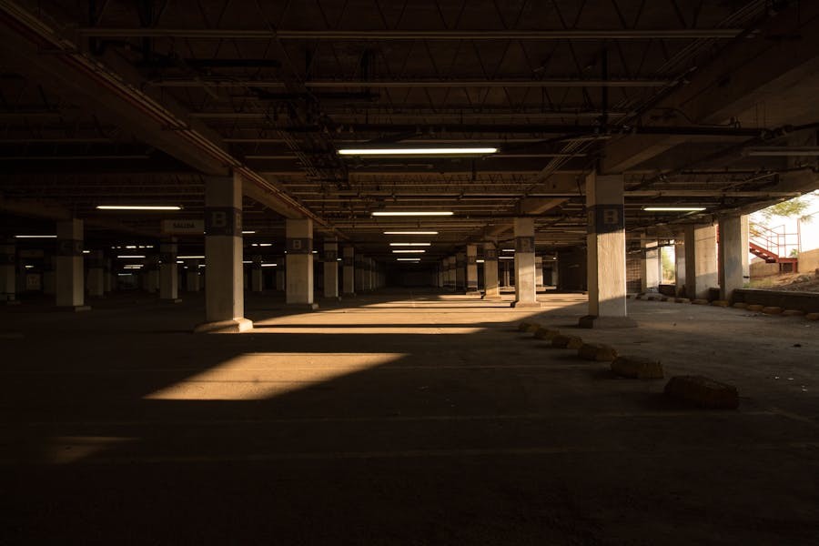 Dimly lit underground parking garage with concrete pillars, overhead fluorescent lighting, and sunlight streaming through an open side entrance.