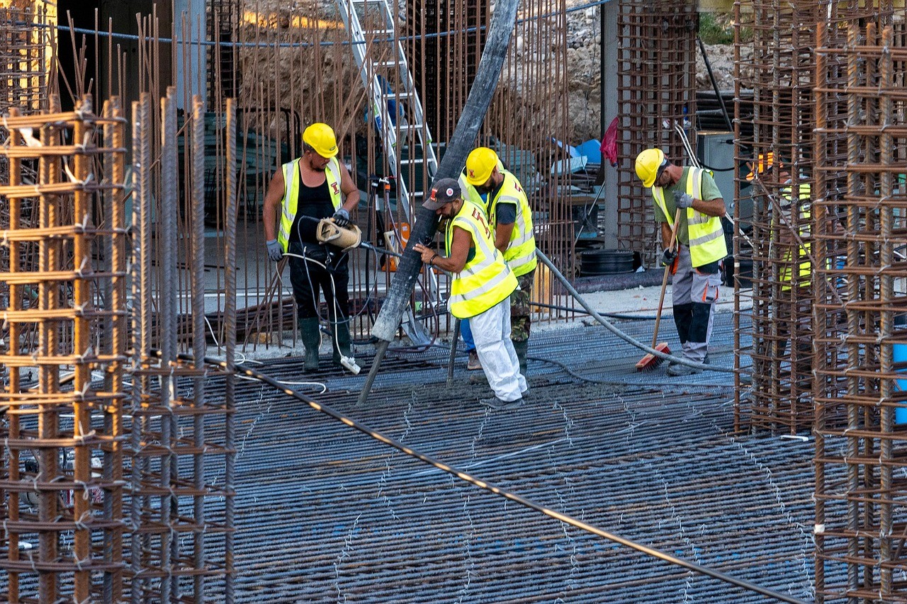 Construction workers in safety gear pouring and spreading concrete over a rebar-reinforced foundation at a building site.