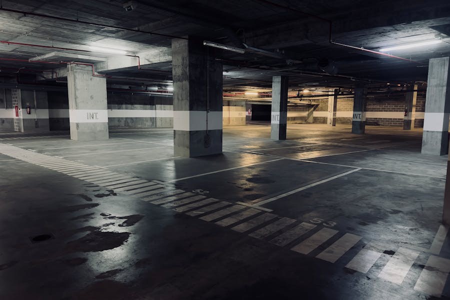 Empty, dimly lit underground parking garage with concrete pillars, marked parking spaces, and overhead lighting casting reflections on the polished floor.