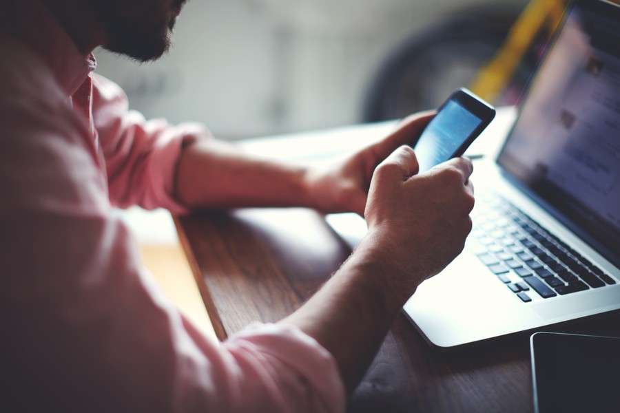 Man holding a phone in front of an open laptop on the desk in front of him.